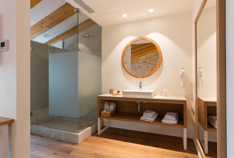 A Forum bathroom: a round oak-framed mirror over a stone basin, a walk-in rain shower behind frosted glass, oak shelving stacked with white towels