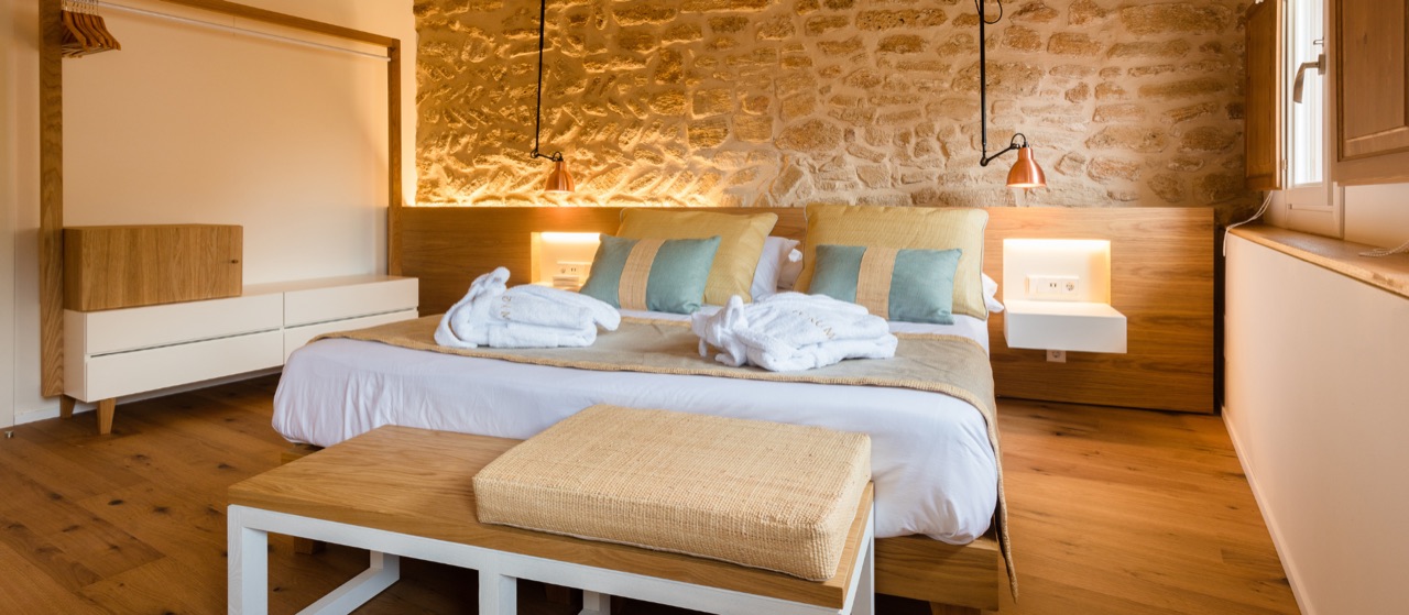 A Forum room with the original rubble-stone wall left bare behind the bed, washed in warm low light, under a ceiling of exposed pine beams