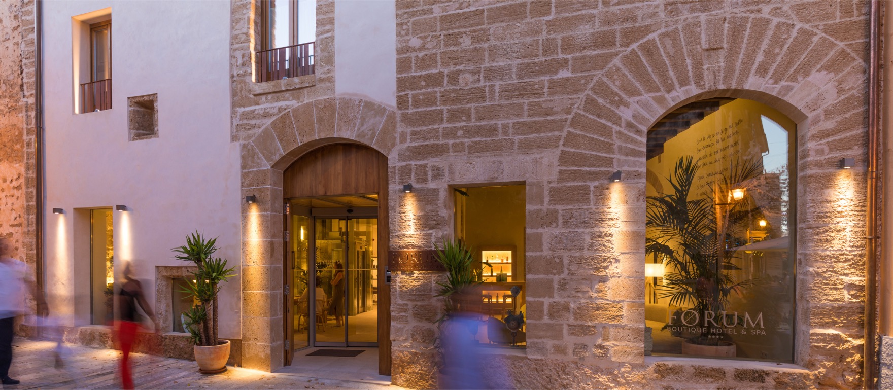 The warm stone entrance of Forum at dusk: a low-lit arched doorway in honey-coloured Alcúdia stone, the carved FORUM sign glowing in a lit window, a narrow old-town street