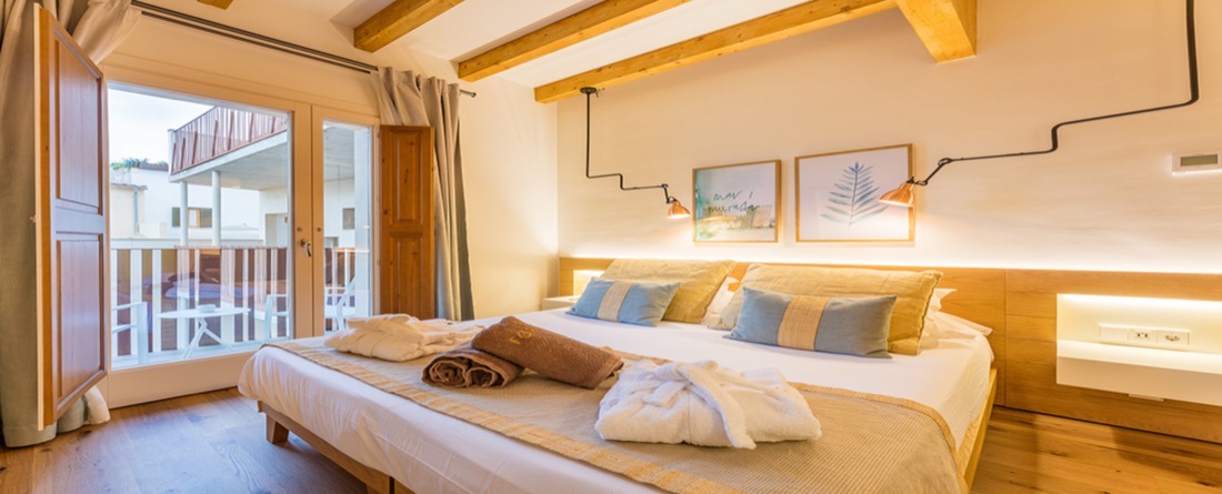 A bright Forum room with beamed ceiling, robes laid on the bed and open shutters onto a small balcony over the Alcúdia rooftops