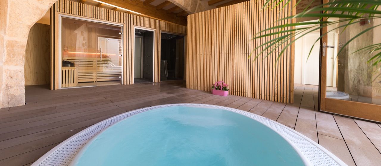 The Forum spa: a round plunge pool of pale blue water set into a warm timber deck, the cedar-lined sauna behind glass and a stone arch to one side
