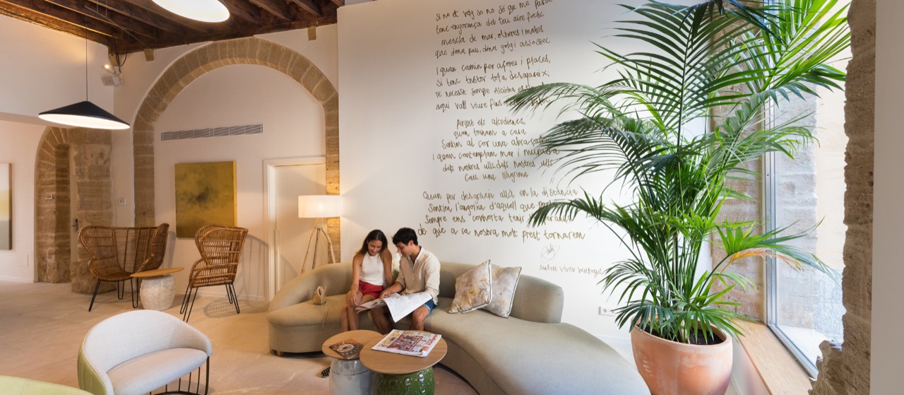The Forum lounge: low cream sofas and woven chairs around a stone arch, a tall palm by the window and a Mallorcan poem written by hand across the white wall